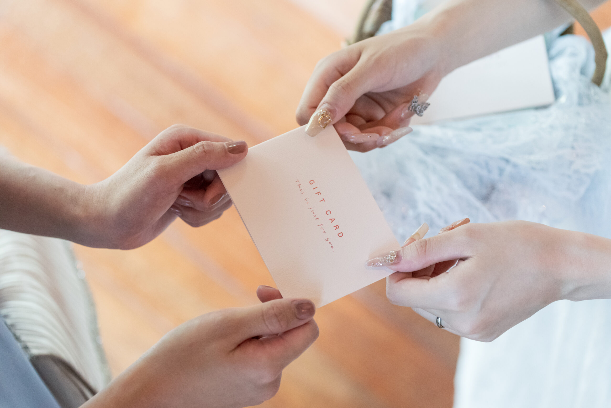 Two pairs of hands exchange a pale pink gift card, with visible text reading 'GIFT CARD' on the card's surface, at a light, pastel-toned setting.