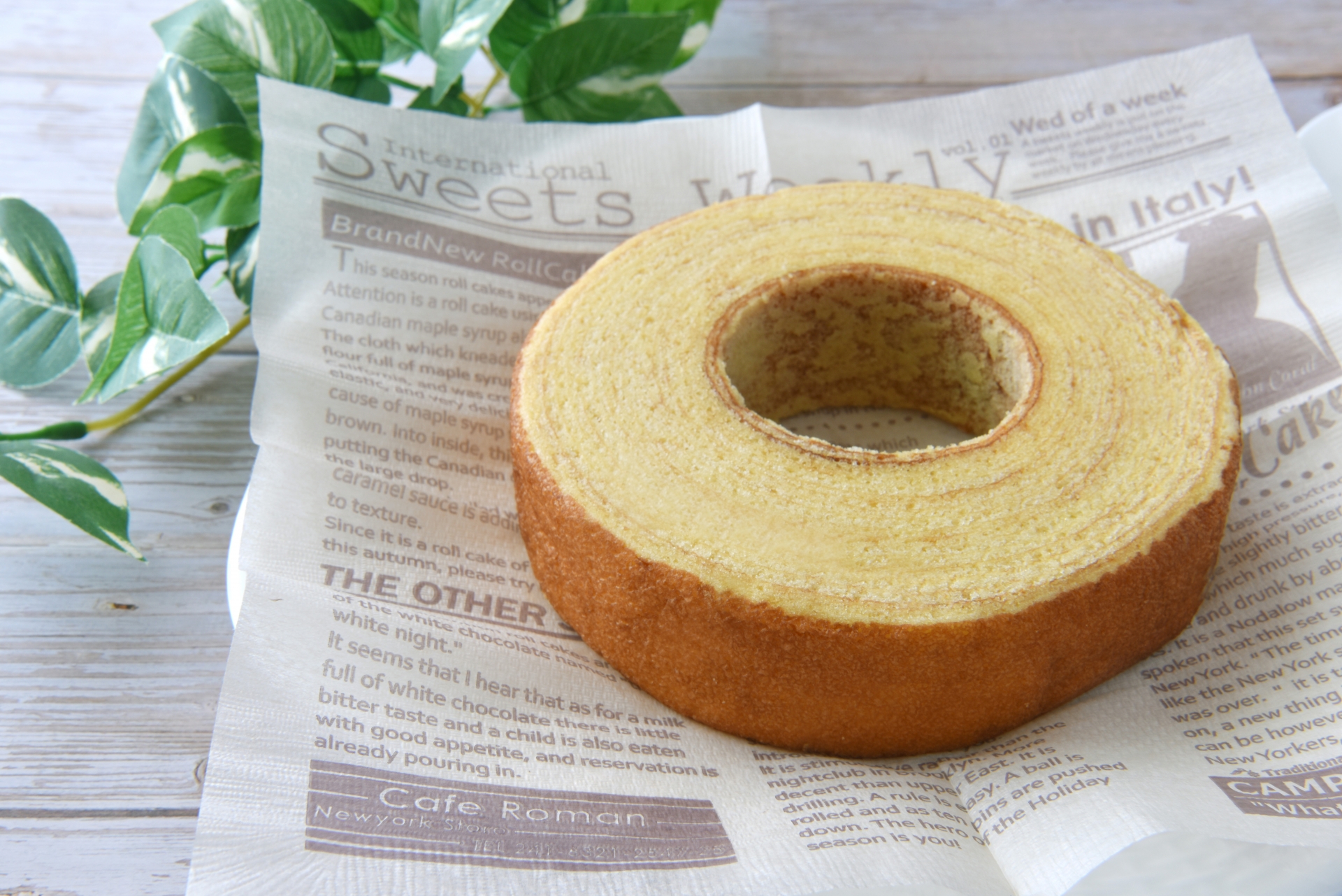 Round vanilla sponge ring cake resting on newspaper-print parchment, with green leaves in the background on a light wooden surface.