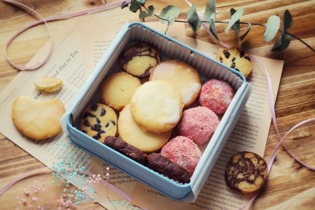 Blue lunchbox filled with assorted cookies on a wooden table, with a paper and eucalyptus nearby.