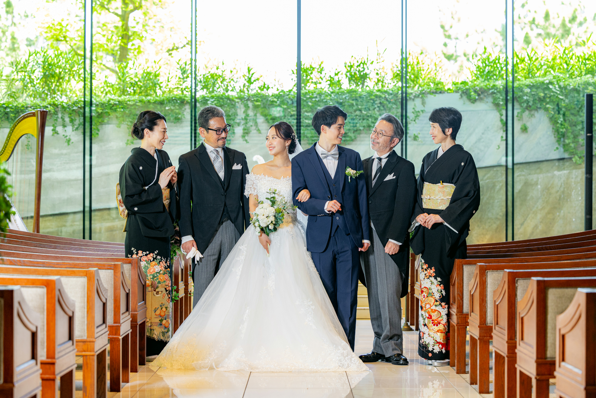 Bride in a white wedding gown and groom in a navy suit walk arm in arm with relatives down a church aisle, smiling together.