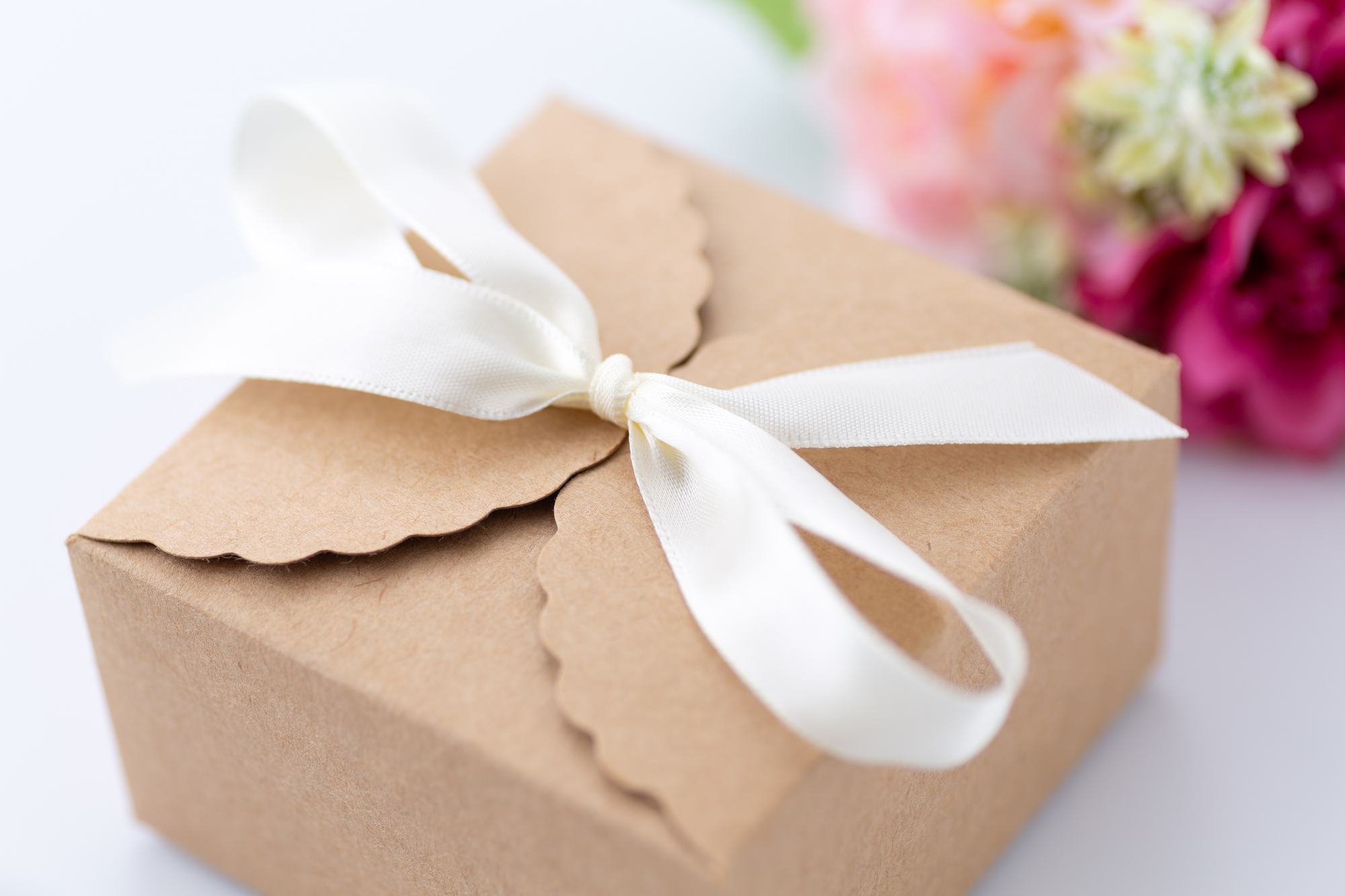 Brown gift box tied with a white ribbon, scalloped lid visible, with colorful flowers in soft focus in the background