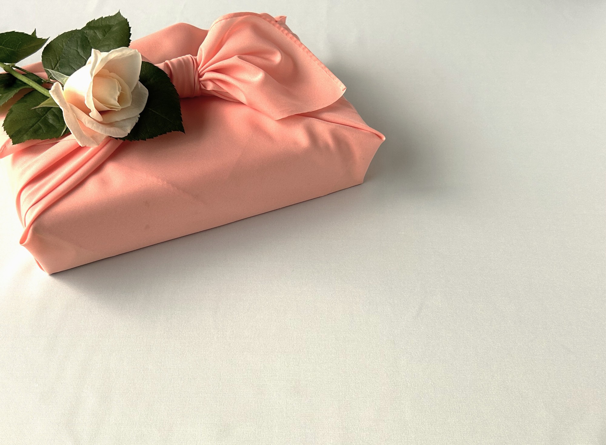 Pink gift box wrapped in satin with a white rose and green leaves on top, resting on a pale surface.